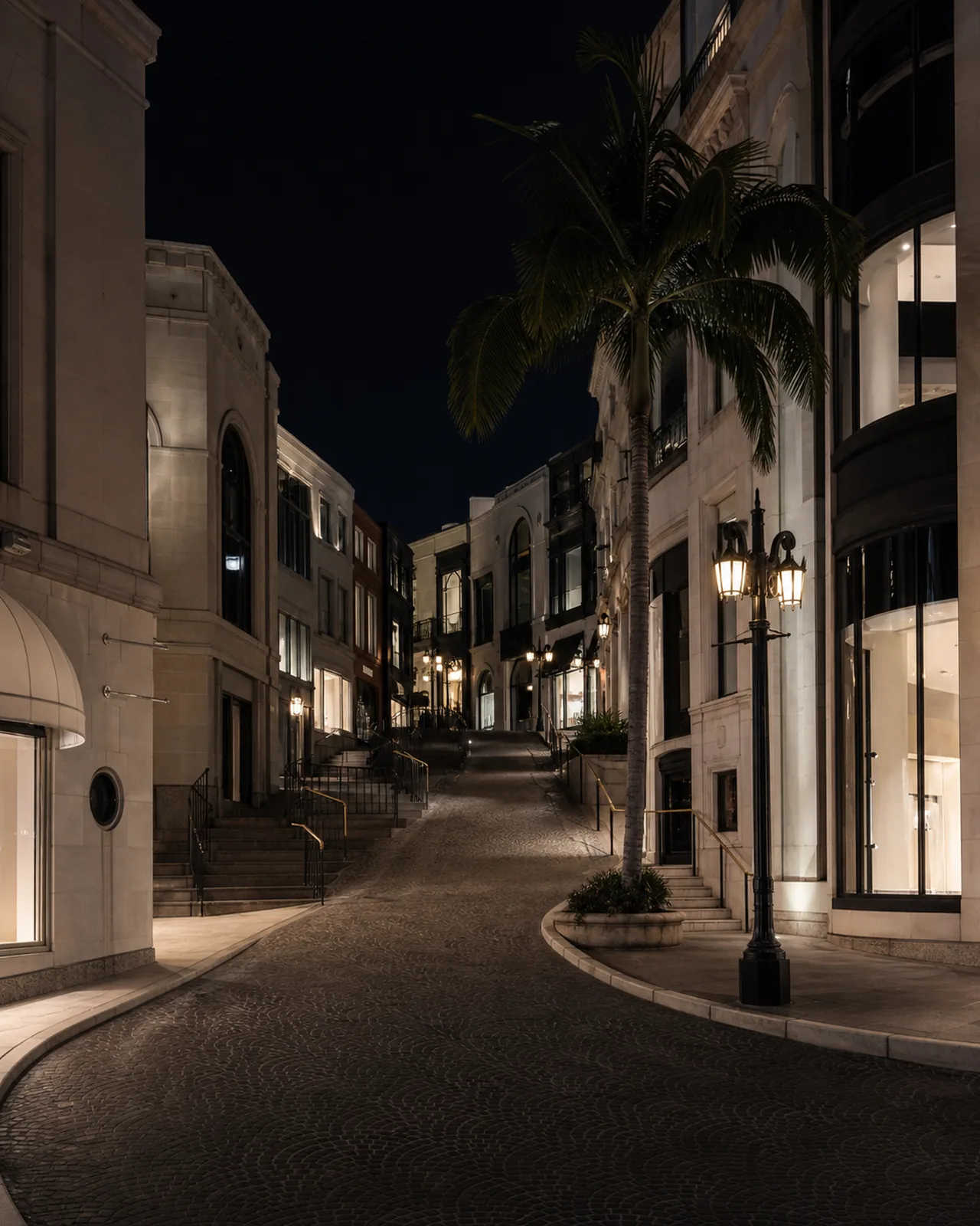 The curved cobblestone Via Rodeo approach lined by limestone storefronts, palms and lamps at blue hour.